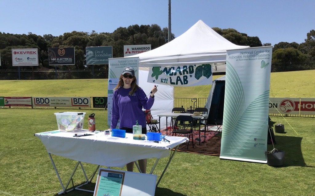 A woman in a purple shirt stands in a grassy oval with a pipette in her hand behind a table of scientific equipment. Behind her is a white tent with a banner reading 'NCARD Lab'.